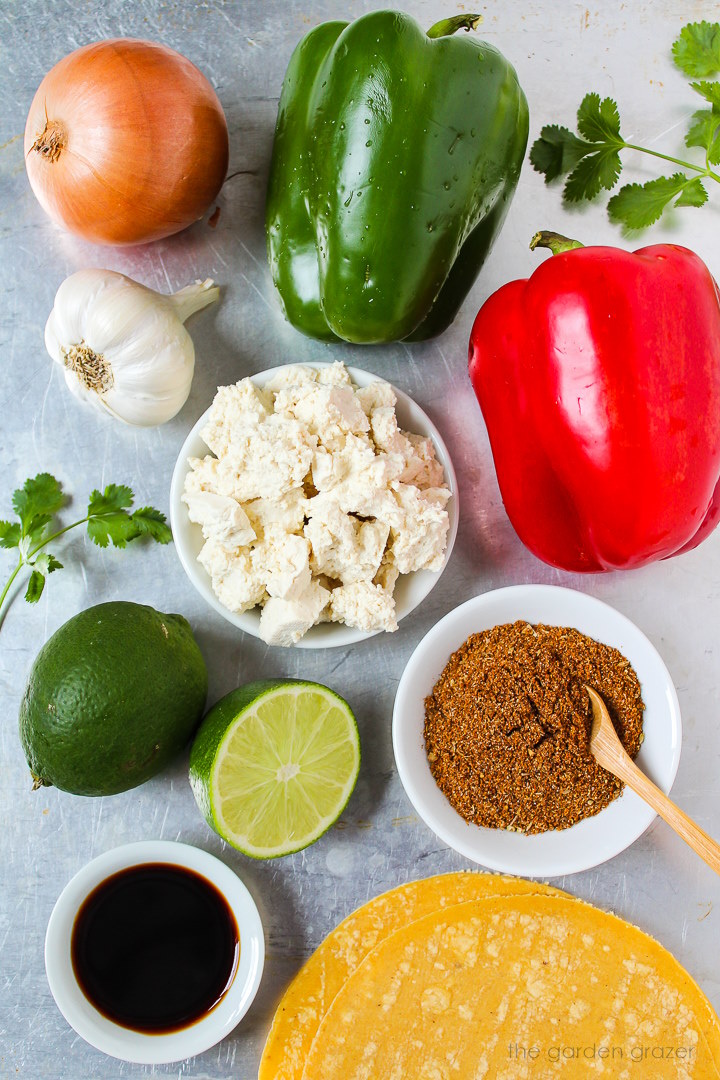 Bell pepper, onion, garlic, corn tortillas, tamari, limes, and seasoning ingredients laid out on a metal tray