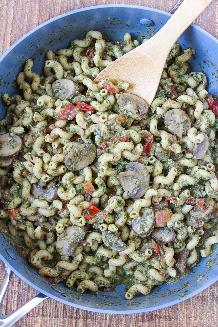 Mushroom pesto pasta cooking in a large skillet with wooden stirring spoon