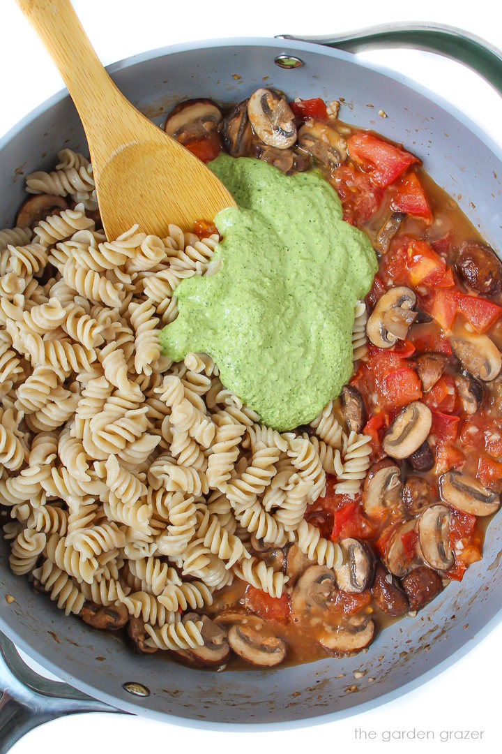 Preparing vegan mushroom pesto pasta in a large skillet with wooden spoon