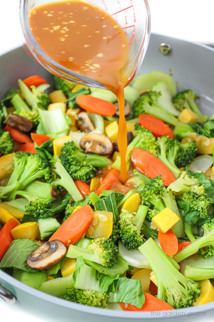 Stir fry sauce being poured into a large pan with cooked vegetables