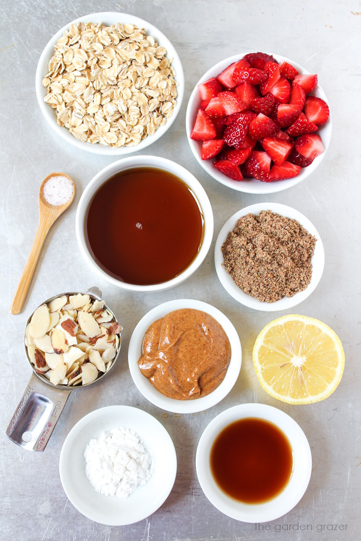 Oats, maple syrup, flax, sliced almonds, almond butter, lemon, and vanilla extract ingredients laid out on a metal tray