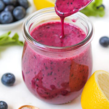 Spoon lifting up blueberry salad dressing from a glass jar on a white table