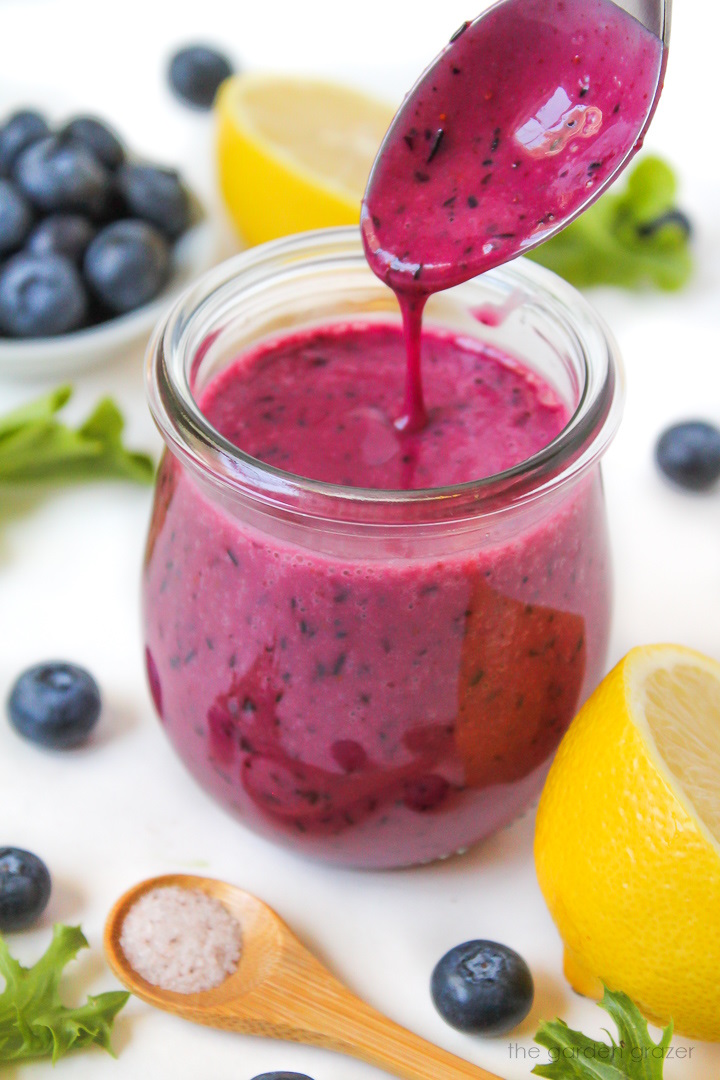 Spoon lifting up blueberry salad dressing from a glass jar on a white table
