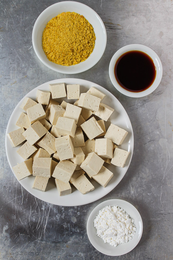 Tofu cubes, nutritional yeast, tamari, and cornstarch ingredients laid out in small white bowls