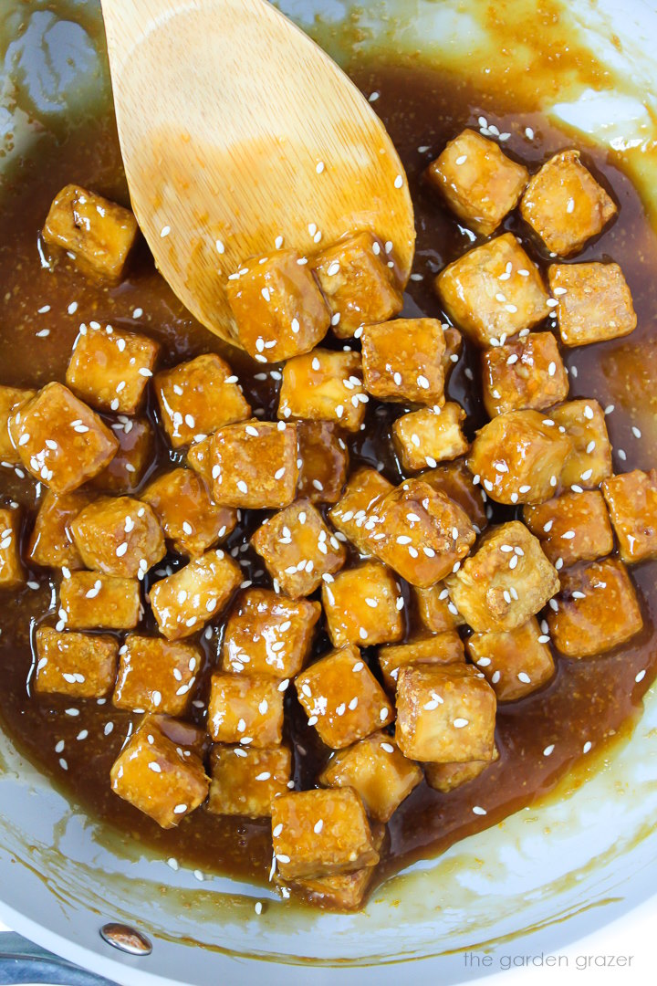 Orange tofu cooking in a skillet with wooden stirring spoon