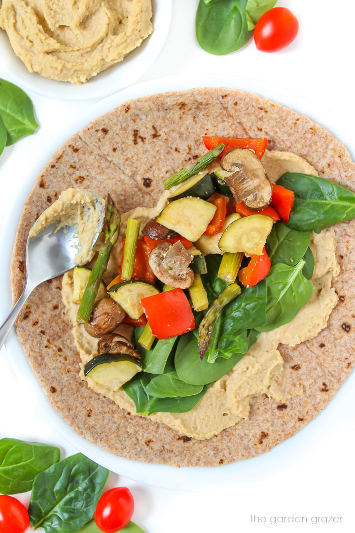 Open-faced roasted veggie wrap with fresh spinach and hummus on a white plate