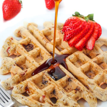 Maple syrup being poured on strawberry waffles on a white plate