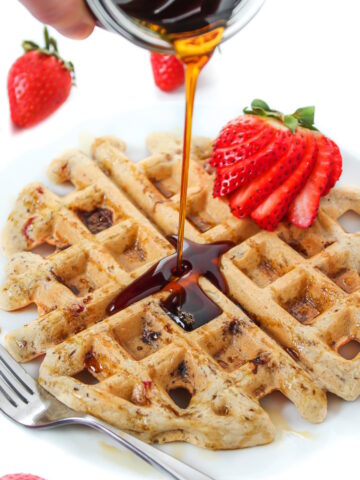 Maple syrup being poured on strawberry waffles on a white plate