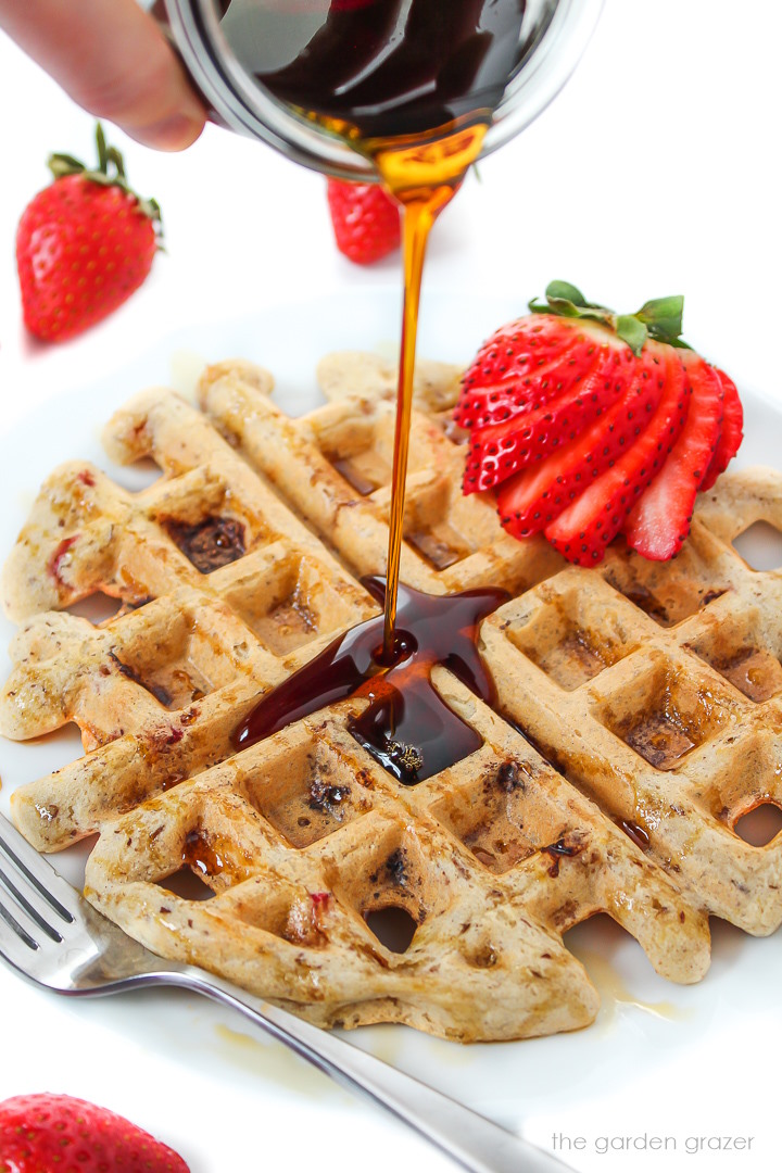 Maple syrup pouring over strawberry waffles on a white plate