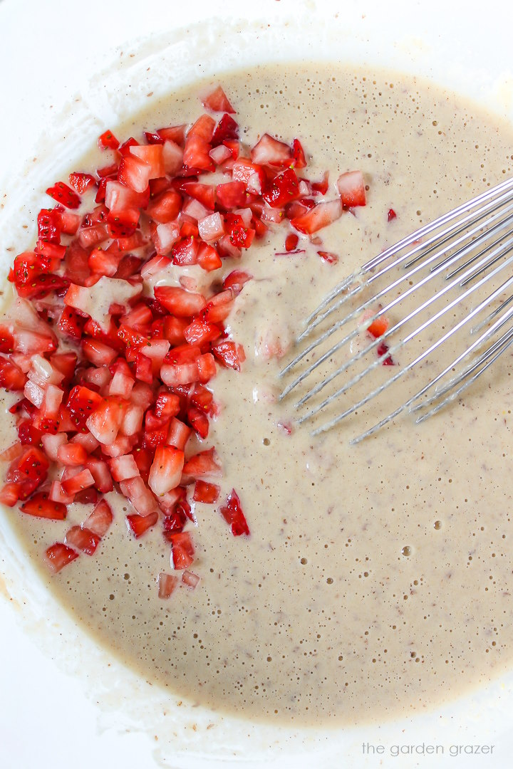 Vegan waffle batter in a large glass bowl with diced strawberries and whisk