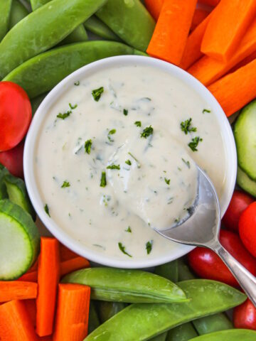 Close up view of vegan ranch dressing in a white bowl with raw veggies for dipping