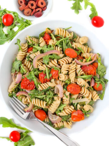 Vegan arugula pasta salad with tomatoes and shallot in a white bowl with serving fork