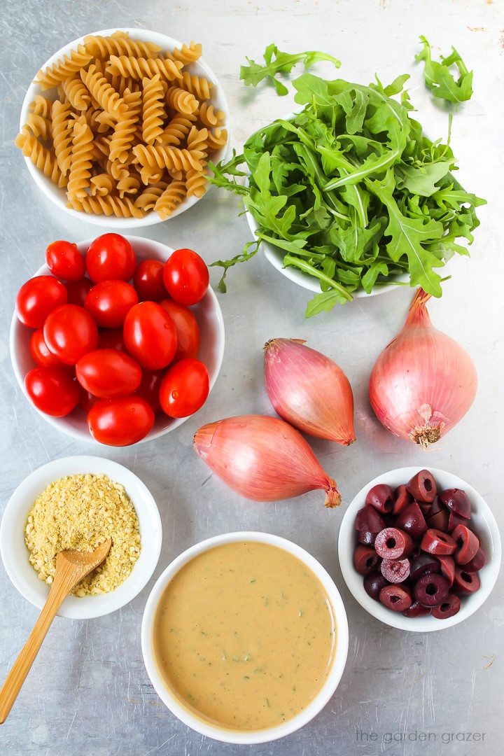 Fresh arugula, fusilli pasta, grape tomatoes, shallots, olives, and Italian dressing ingredients laid out on a metal tray
