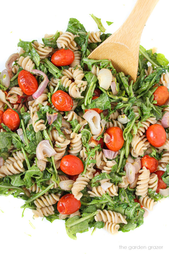Tossing together arugula pasta salad with tomatoes and shallots in a large glass bowl