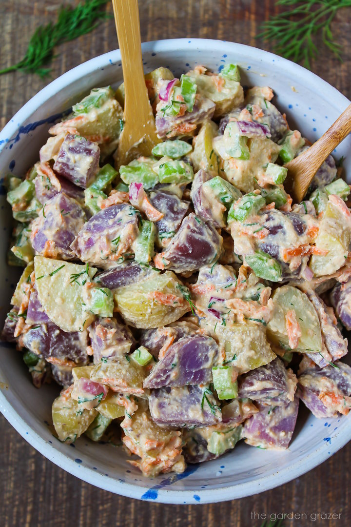 Vegan potato salad with celery, carrots, and onion in a white bowl with serving spoons