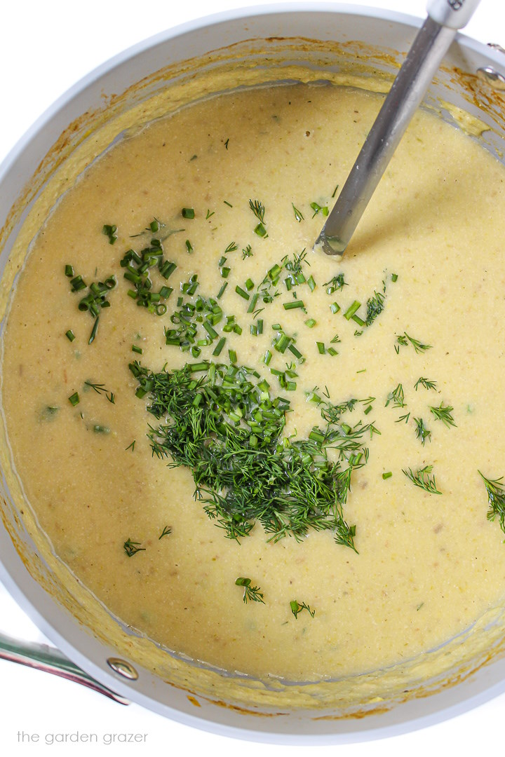Blended vegan potato and leek soup topped with fresh herbs in a large stockpot with metal ladle