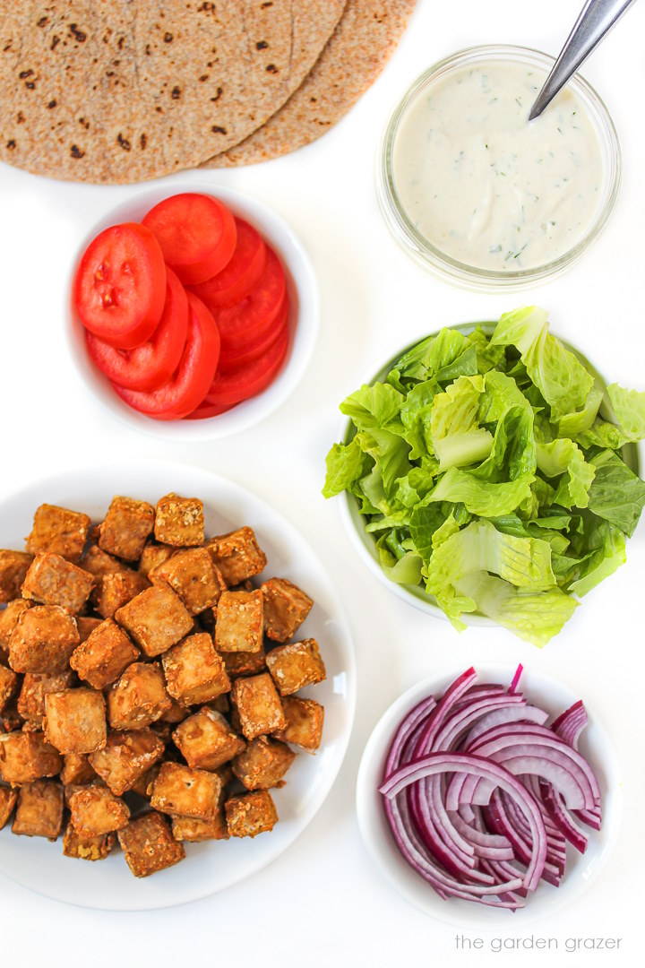 Tortillas, tomatoes, lettuce, dressing, and onion ingredients laid out on a white table