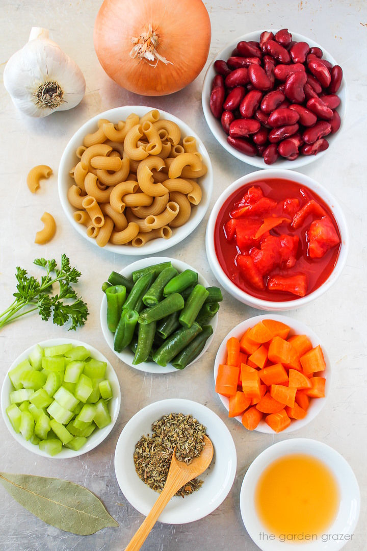 Overhead view of elbow pasta, diced tomatoes, kidney beans, onion, garlic, carrot, celery, and seasoning ingredients laid out on a metal tray