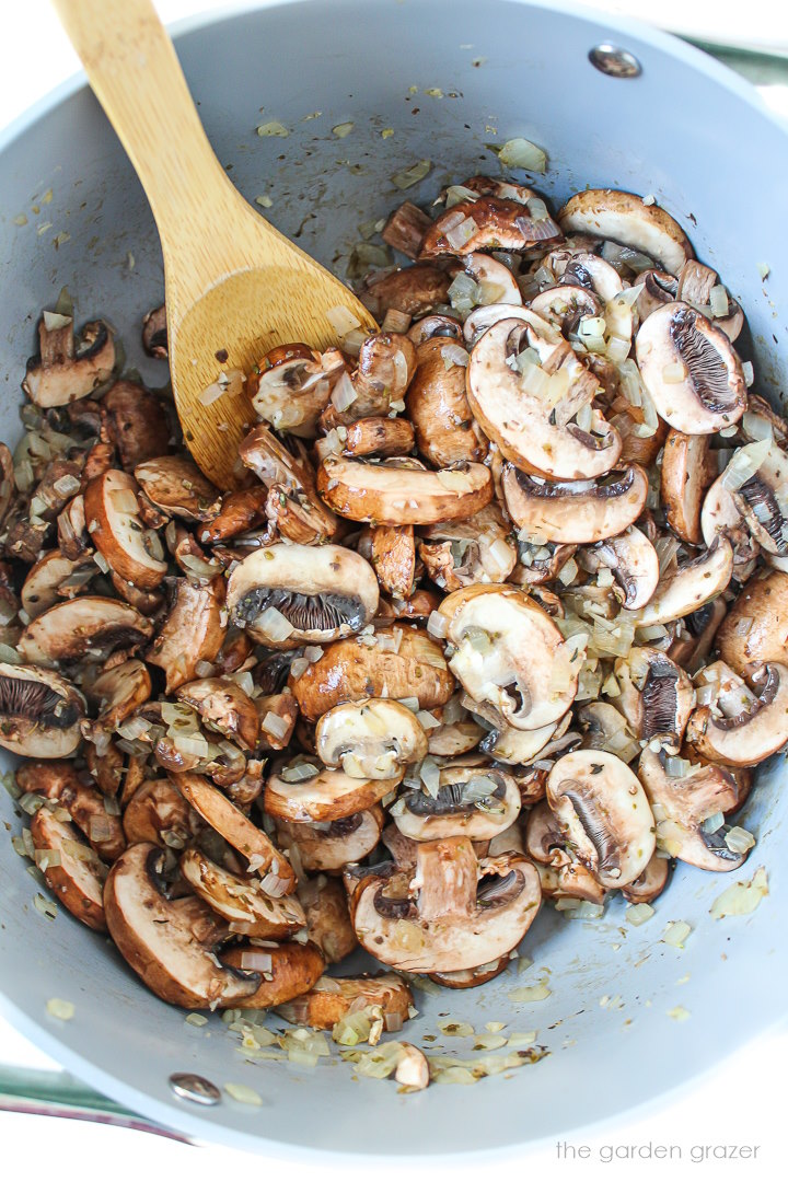 Mushrooms, onions, and herbs cooking in a pot with wooden stirring spoon