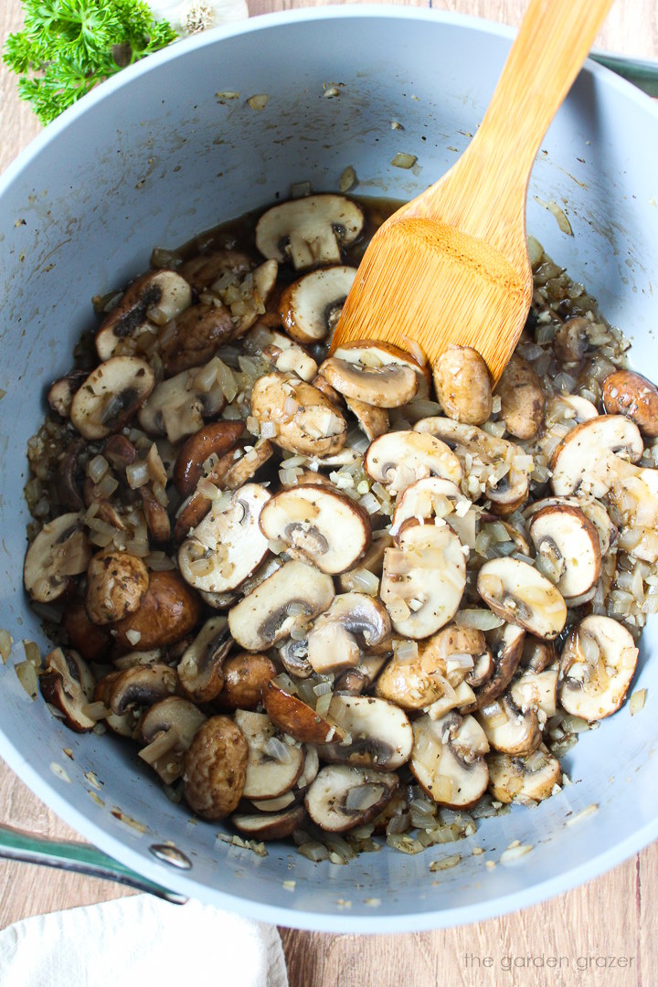 Mushrooms, onions, and herbs cooking in a pot with wooden stirring spoon