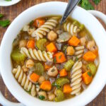 Chickpea noodle soup in a white bowl with serving spoon sitting on a wooden table