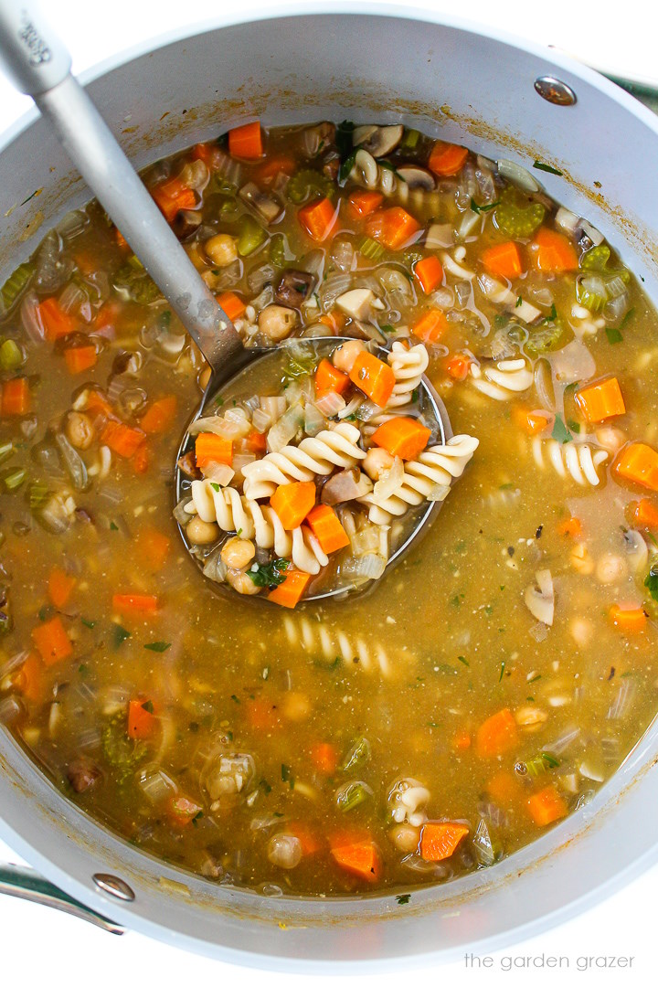 Overhead view of a metal ladle lifting up chickpea noodle soup as it cooks in a large pot