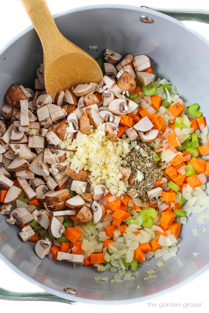 Overhead view of preparing onions, celery, carrot, garlic, and herbs in a large pot with stirring spoon