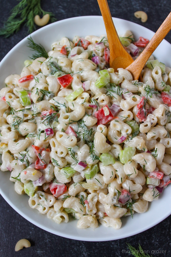 Vegan macaroni salad with dill in a white serving bowl with wooden spoons