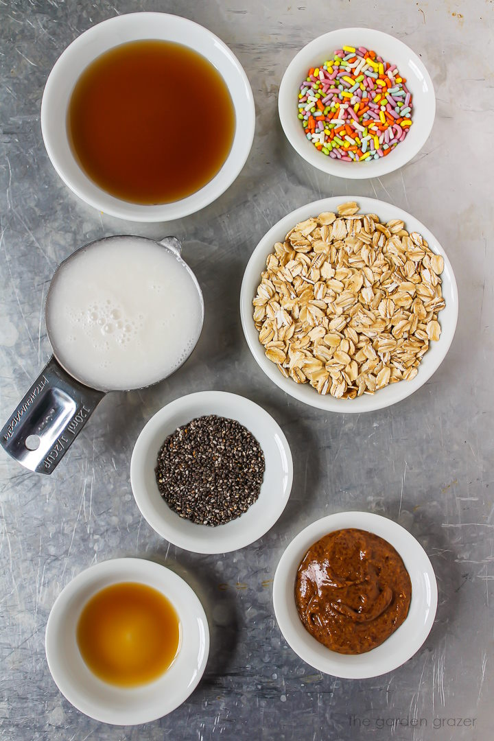 Milk, oats, maple syrup, chia seeds, sprinkles, and vanilla extract ingredients laid out on a metal tray