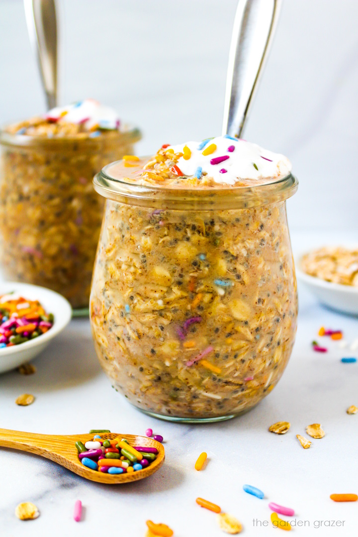Two glass jars of prepared birthday cake overnight oats with whipped cream and rainbow sprinkles