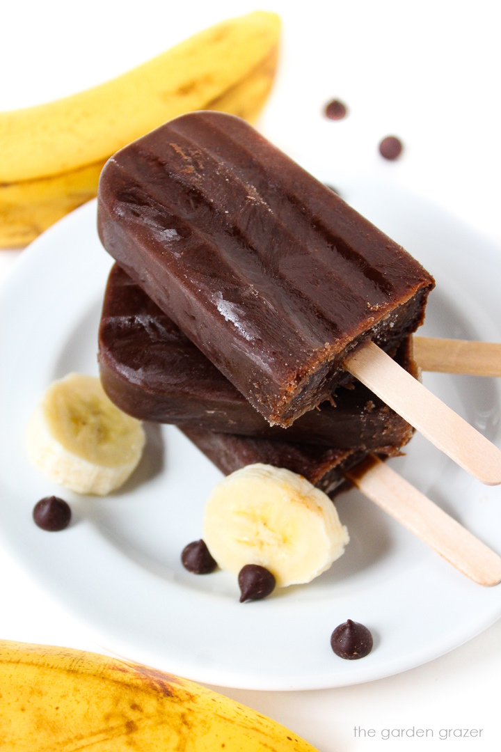 Three chocolate banana popsicles stacked on a white plate with banana slices