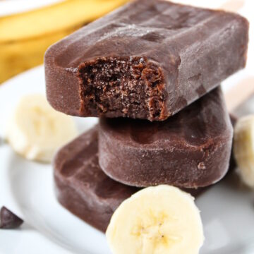 Three vegan chocolate banana popsicles stacked on a plate with a bite taken out of one