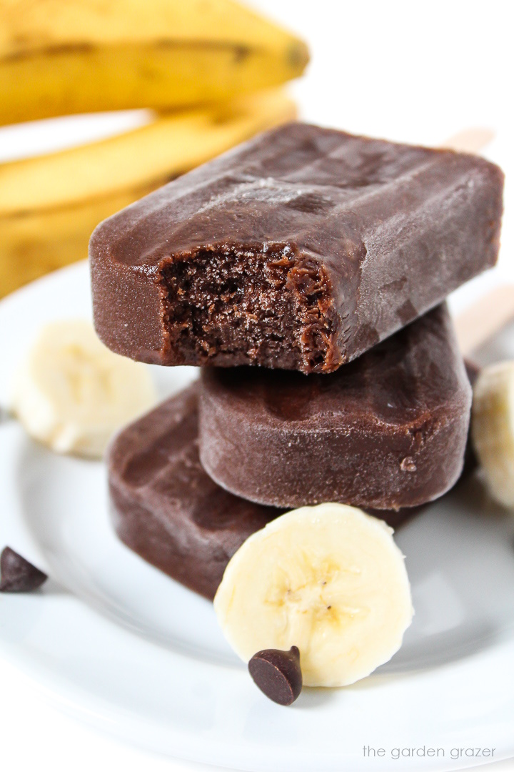 Three vegan chocolate banana popsicles stacked on a plate with a bite taken out of one