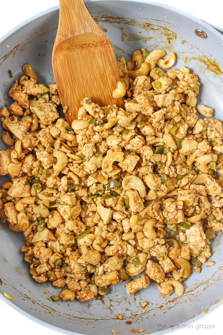 Cashew tofu mixture with savory sauce cooking in a skillet with wooden spatula