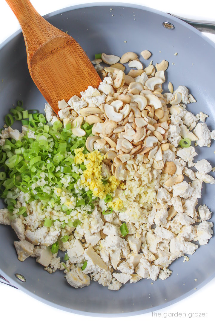 Crumbled tofu, green onions, garlic, and ginger cooking in a skillet with stirring spoon