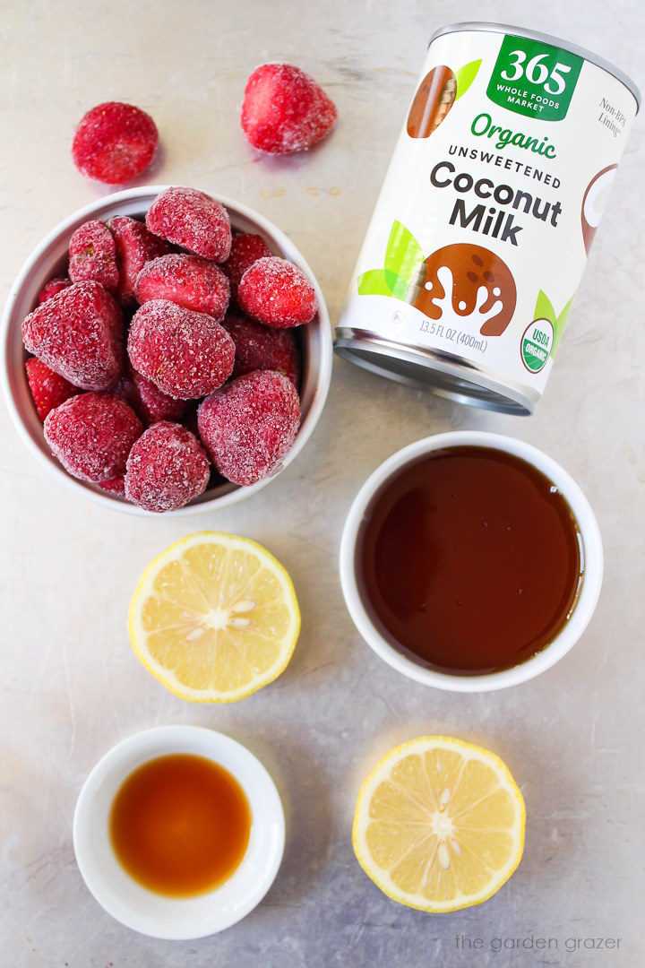 Frozen strawberries, coconut milk, vanilla extract, lemon, and maple syrup ingredients laid out on a metal tray