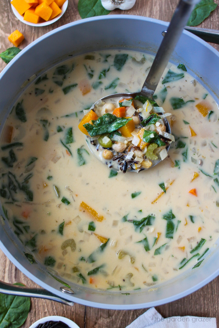 A metal ladle lifting up creamy butternut squash wild rice soup out of a pot after it cooks