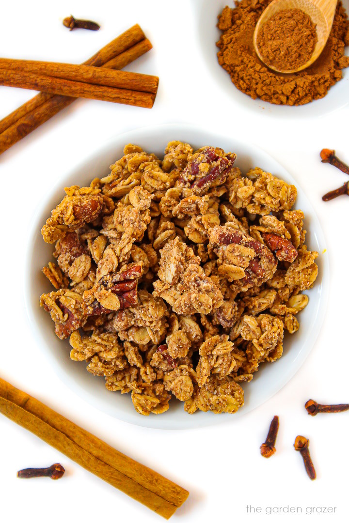 Vegan pumpkin spice granola in a white bowl surrounded with cinnamon and cloves