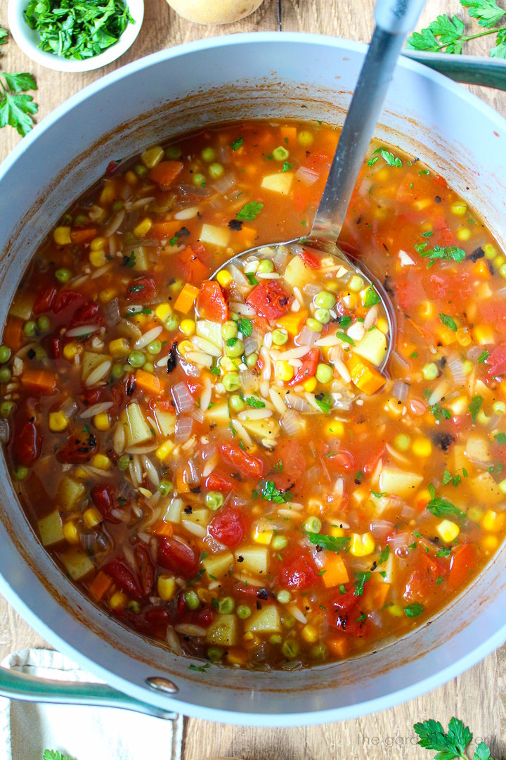 Overhead view of a ladle scooping out vegetable orzo soup from a large pot as it's cooking