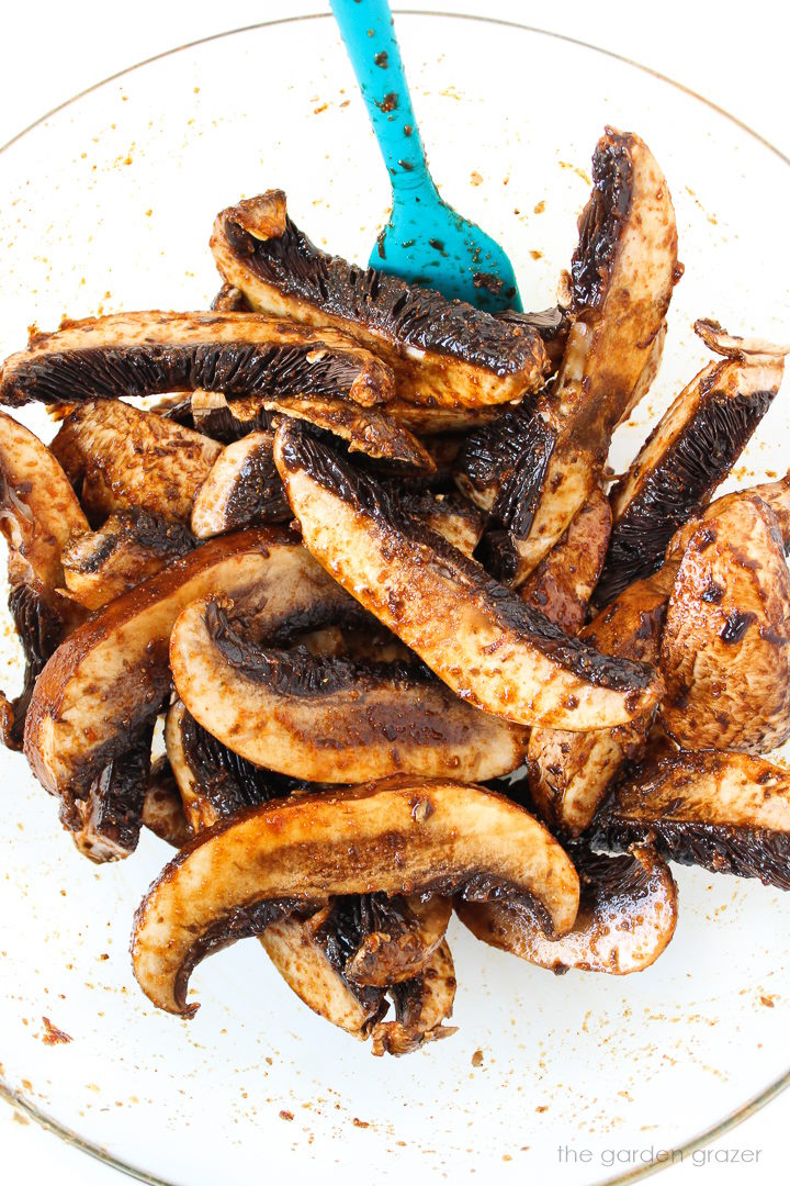 Overhead view of sliced portabella mushrooms in a large glass bowl tossed in a savory marinade sauce before roasting