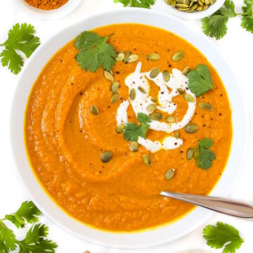 Overhead view of vegan pumpkin curry soup in a white bowl topped with yogurt, pumpkin seeds, and fresh cilantro