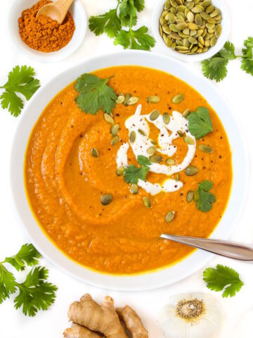 Overhead view of vegan pumpkin curry soup in a white bowl topped with yogurt, pumpkin seeds, and fresh cilantro