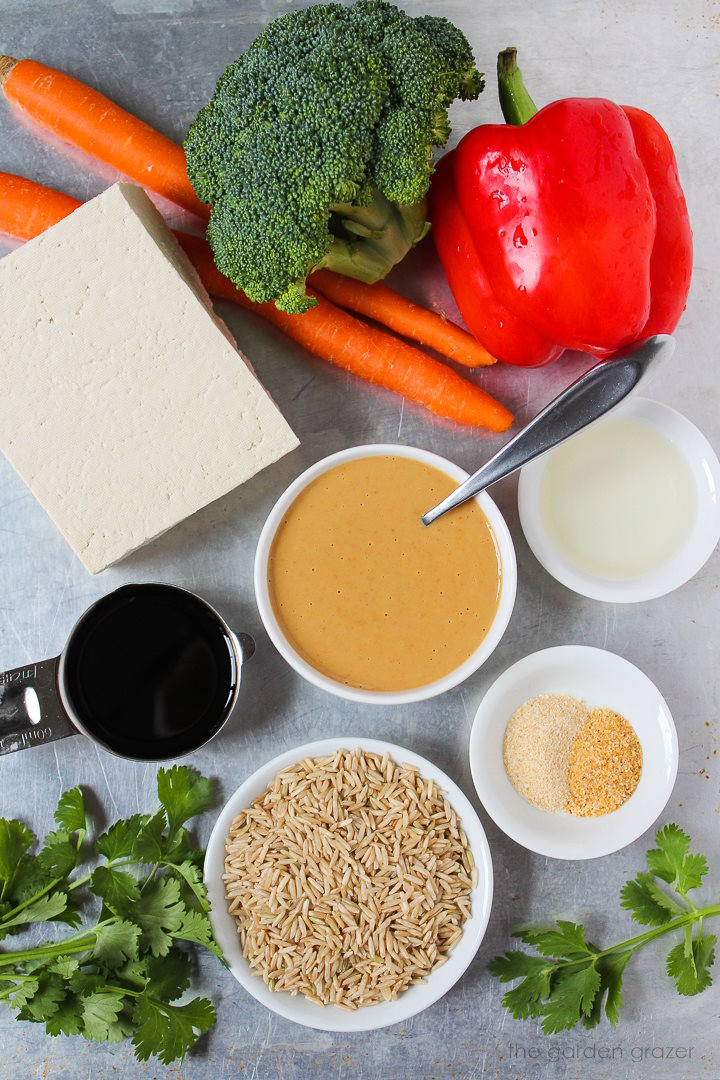 Fresh broccoli, carrots, bell pepper, tofu, rice, peanut sauce, and spice ingredients laid out on a metal tray