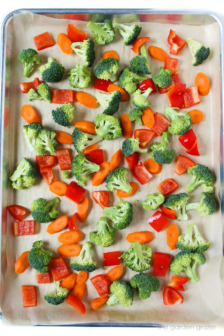Fresh broccoli florets, sliced carrots, and chopped bell pepper spread out on a sheet pan before roasting