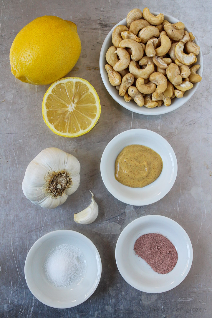 Raw cashews, fresh lemon, garlic bulb, mustard, and salt ingredients laid out on a metal tray