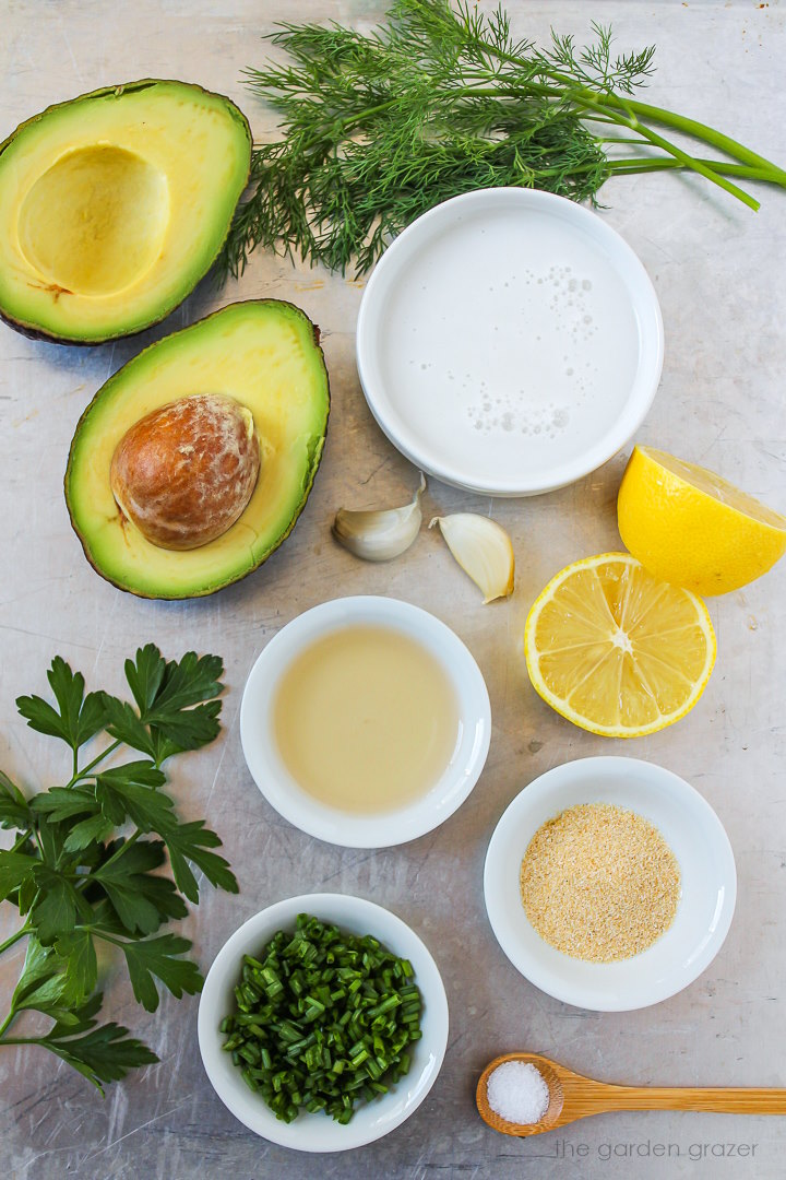 Avocado, lemon, dairy-free milk, vinegar, garlic, and fresh herb ingredients laid out on a metal tray