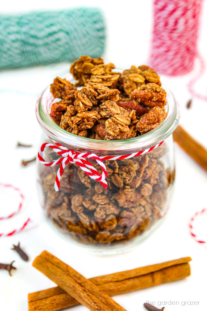 Vegan gingerbread granola in a small glass jar with cinnamon sticks and cloves on the side