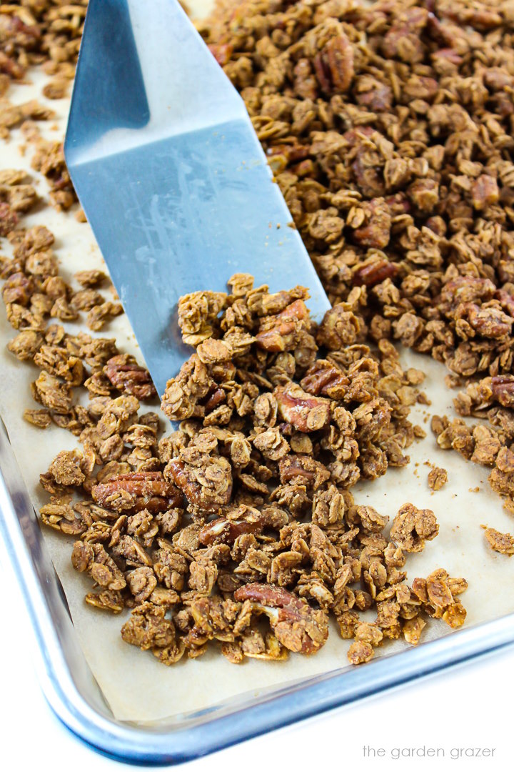 Metal spatula scooping up gingerbread granola from a sheet pan