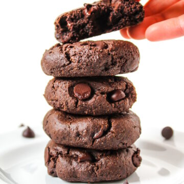 A hand taking the top vegan chocolate cookie from a stack on a white plate