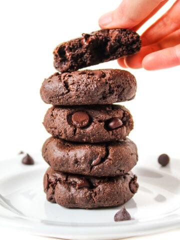 A hand taking the top vegan chocolate cookie from a stack on a white plate
